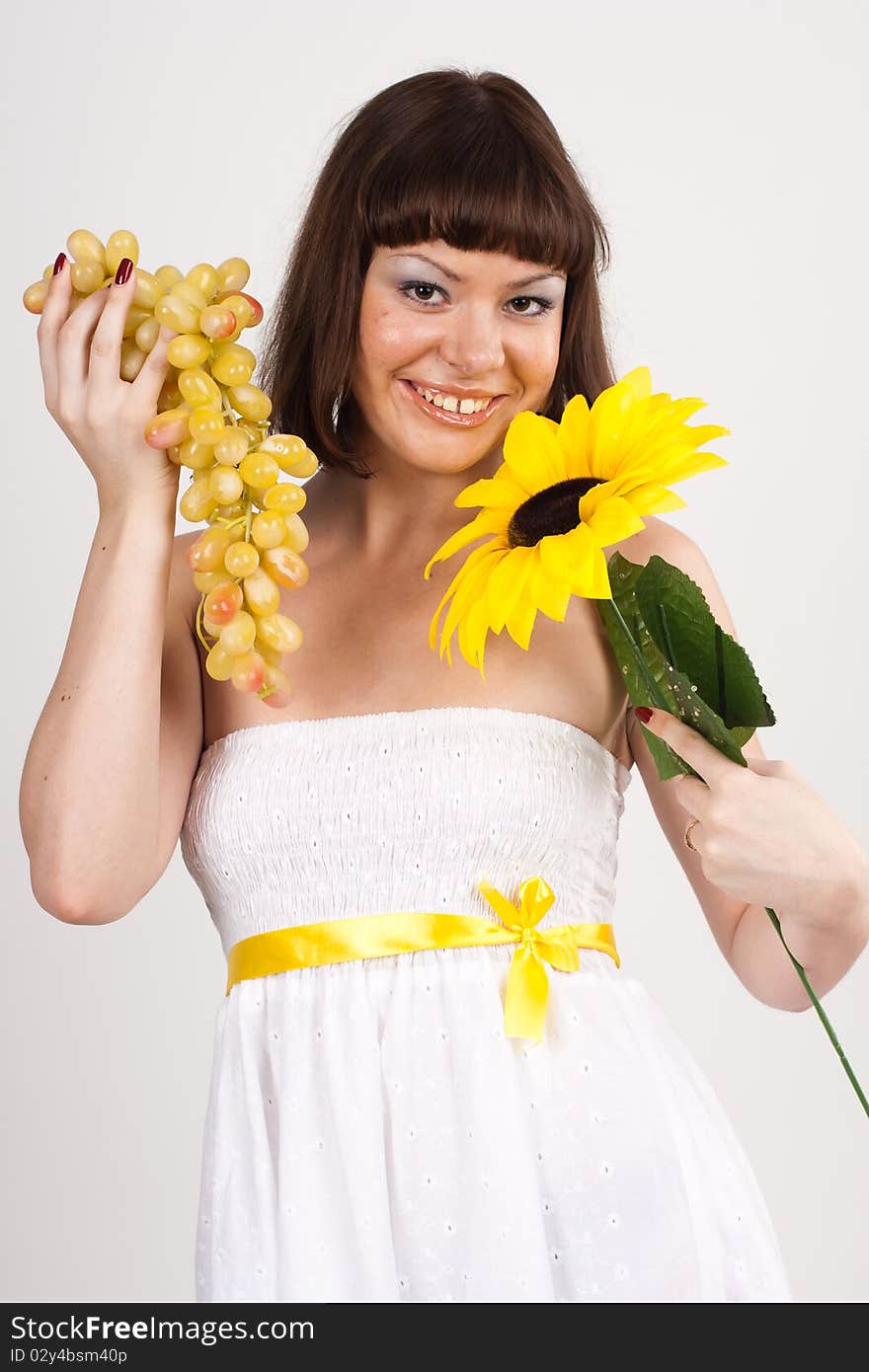 Beautiful girl in white dress with yellow tape is holding green grapes and sunflower. She is isolated on a white background. Beautiful girl in white dress with yellow tape is holding green grapes and sunflower. She is isolated on a white background.