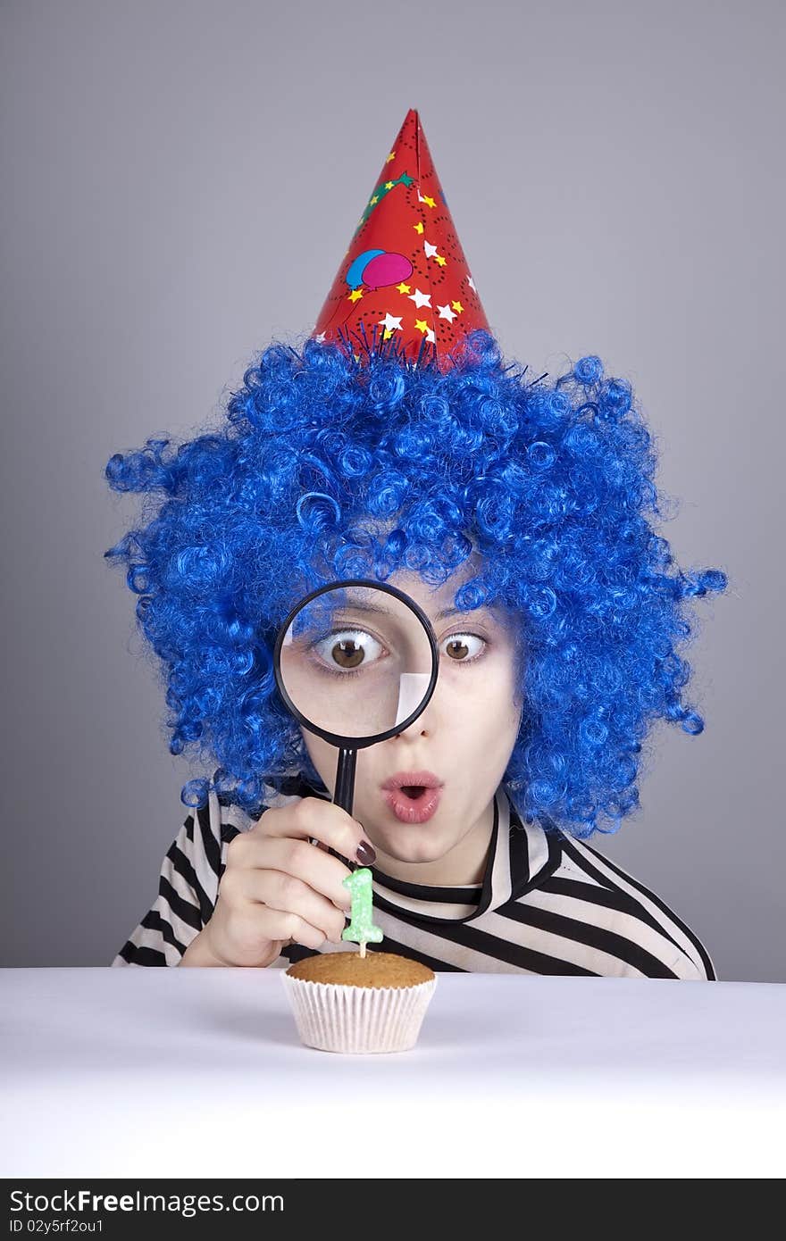 Funny girl with blue hair and striped knitted jacket looking through loupe at cake for birthday with one candle. Funny girl with blue hair and striped knitted jacket looking through loupe at cake for birthday with one candle.