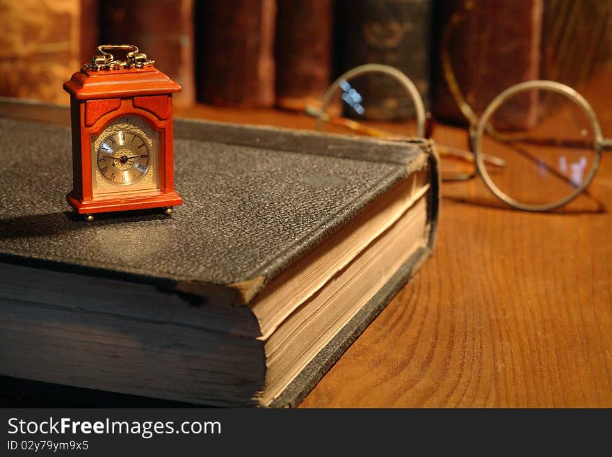 Vintage still life. Small wooden clock standing on old book on background with books and spectacles