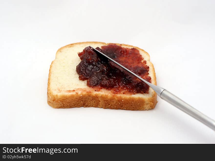 A piece of white bread with strawberry jelly being spread over it with a butter knife. A piece of white bread with strawberry jelly being spread over it with a butter knife.