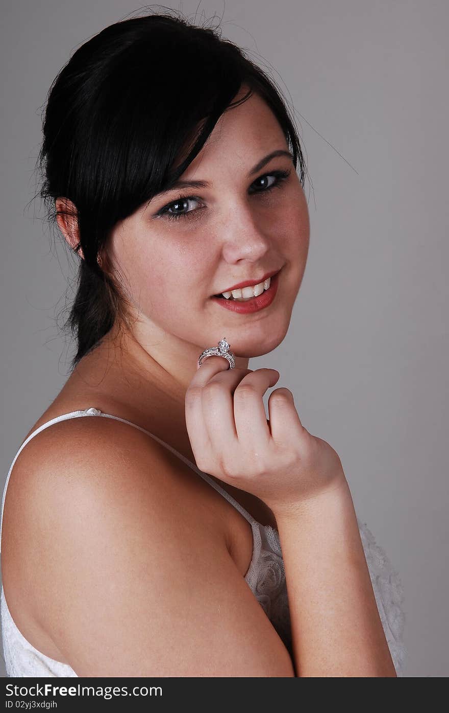 A beautiful young woman holds up her new engagement diamond ring, 
head shot over light gray background. A beautiful young woman holds up her new engagement diamond ring, 
head shot over light gray background.