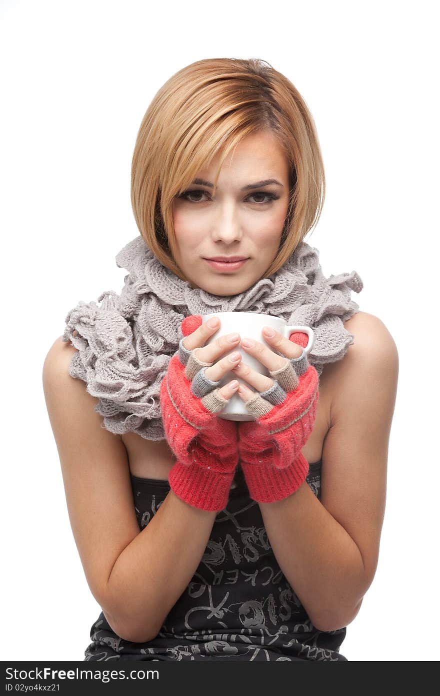 A frontal portrait of a young, blonde woman, holding a mug with both her hands, wearing a muffler and winter gloves, with an expression of being cold. A frontal portrait of a young, blonde woman, holding a mug with both her hands, wearing a muffler and winter gloves, with an expression of being cold.