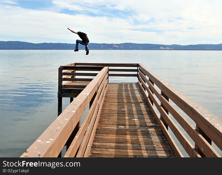 A man jumping a long narrow wooden pier into the calm blue ocean water with outstretched victorious arms. A man jumping a long narrow wooden pier into the calm blue ocean water with outstretched victorious arms.