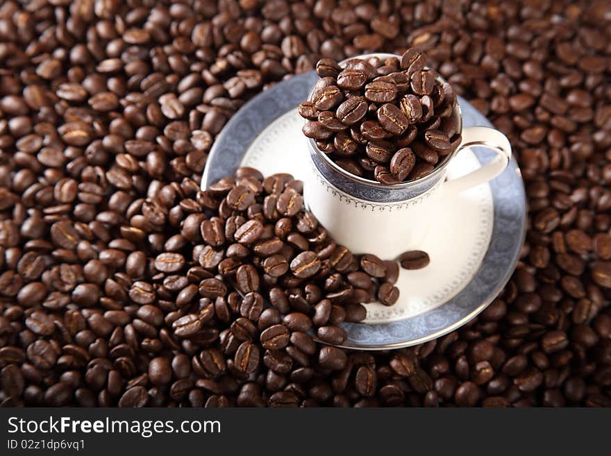 Cup and saucer filled to the top with roasted coffee beans placed on a background of coffee beans. Cup and saucer filled to the top with roasted coffee beans placed on a background of coffee beans