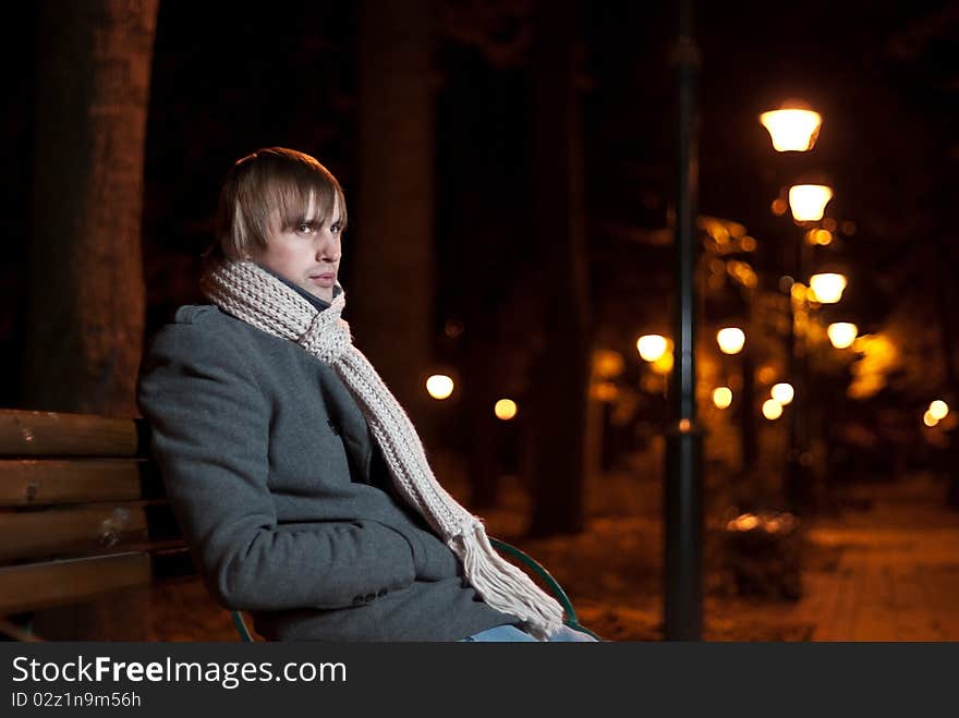 Young man in coat sitting at night park alley. Young man in coat sitting at night park alley