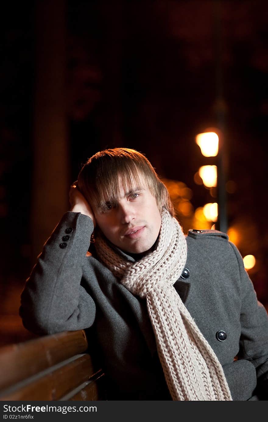 Young man in coat sitting at night park alley. Young man in coat sitting at night park alley