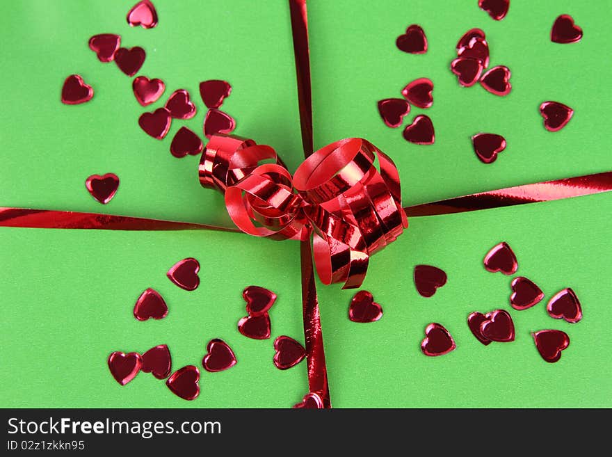 Gift in green wrapping with a red bow decorated with heart shaped confetti in close up. Gift in green wrapping with a red bow decorated with heart shaped confetti in close up