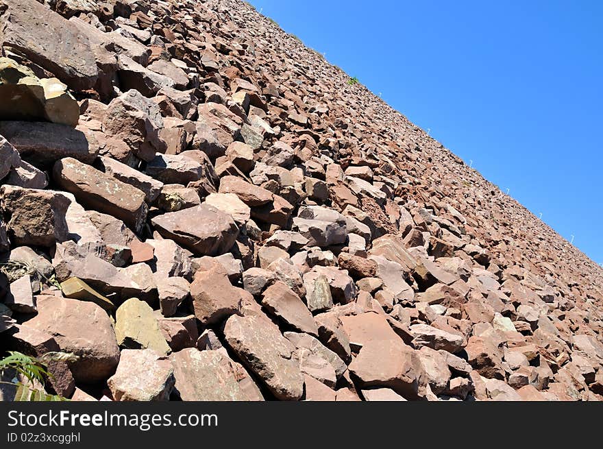 Slope of water dam, construction area full of stone in brownness, under blue sky, shown as perspective and industrial solid and powerful. Slope of water dam, construction area full of stone in brownness, under blue sky, shown as perspective and industrial solid and powerful.