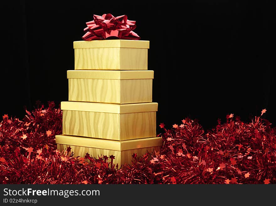 Four gold boxes stacked with a red bow on top one and sitting on red tinsel garland. Four gold boxes stacked with a red bow on top one and sitting on red tinsel garland