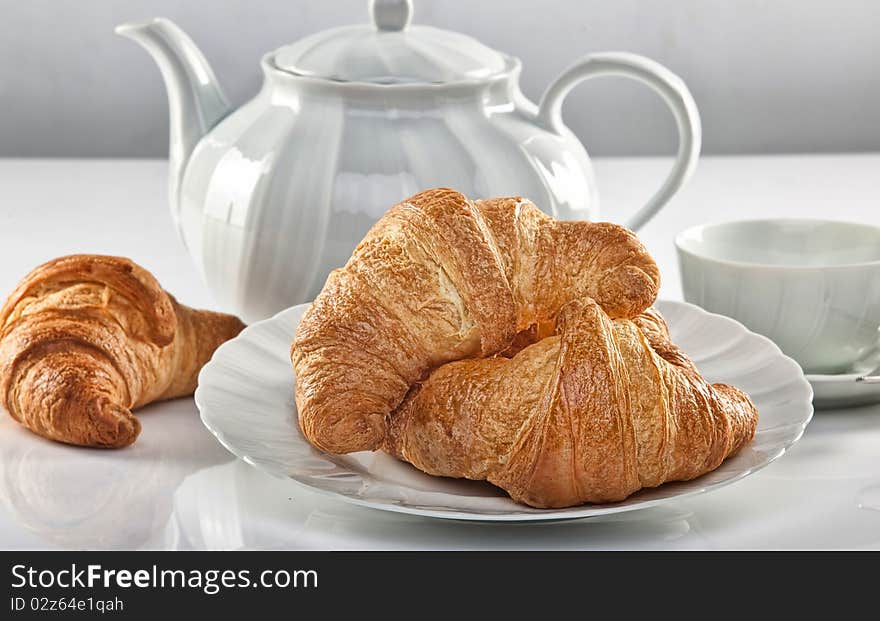 It's time for breakfast! Some fragrant croissant on a white dish. A tea pot is on the background. It's time for breakfast! Some fragrant croissant on a white dish. A tea pot is on the background