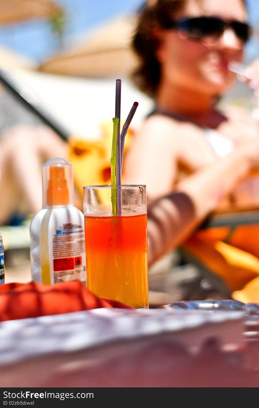 Cocktail glass with two straws, a young women relaxing in the brackground and a sunscreen