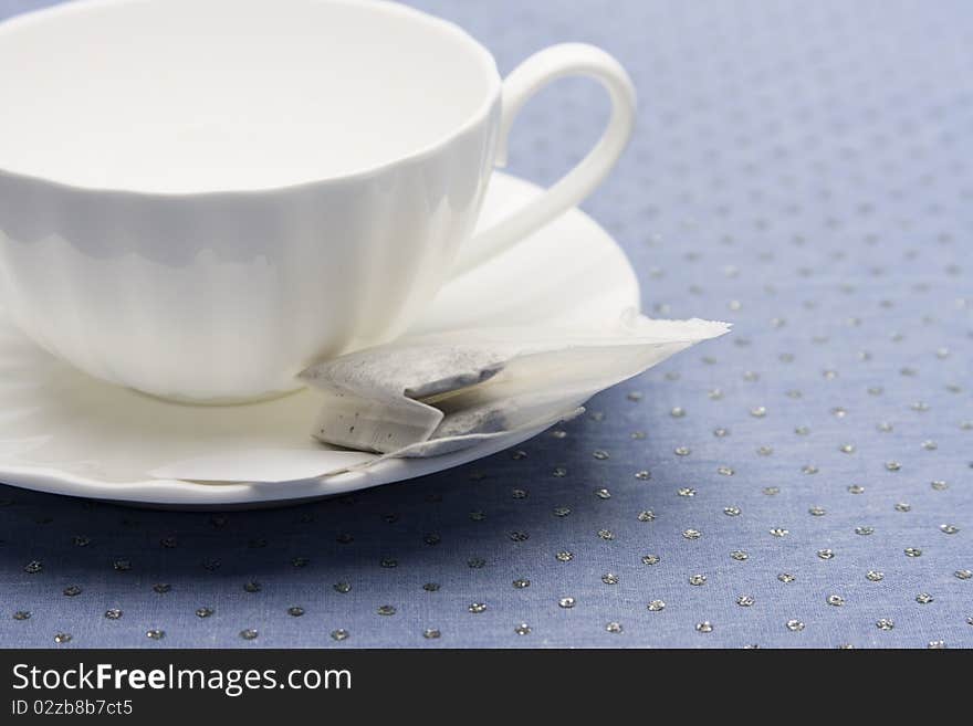 Empty white china tea cup and saucer with tea bag laying on top of a glitter blue tabletop.