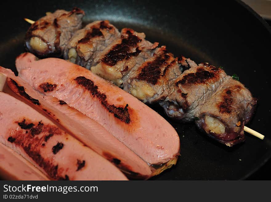 The image depicts a plate of mixed meat, kebabs and wustel during cooking, in a frying pan.