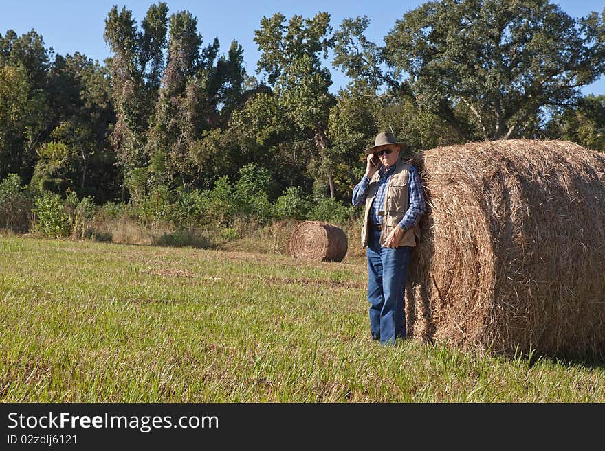 Mature man wearing hat and vest, standing next to a bale of hay and talking on cell phone. Mature man wearing hat and vest, standing next to a bale of hay and talking on cell phone.