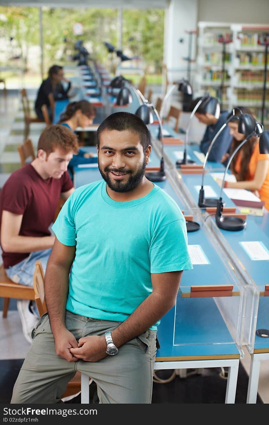 Portrait of male college student sitting on table in library and looking at camera. Vertical shape, high angle view. Portrait of male college student sitting on table in library and looking at camera. Vertical shape, high angle view