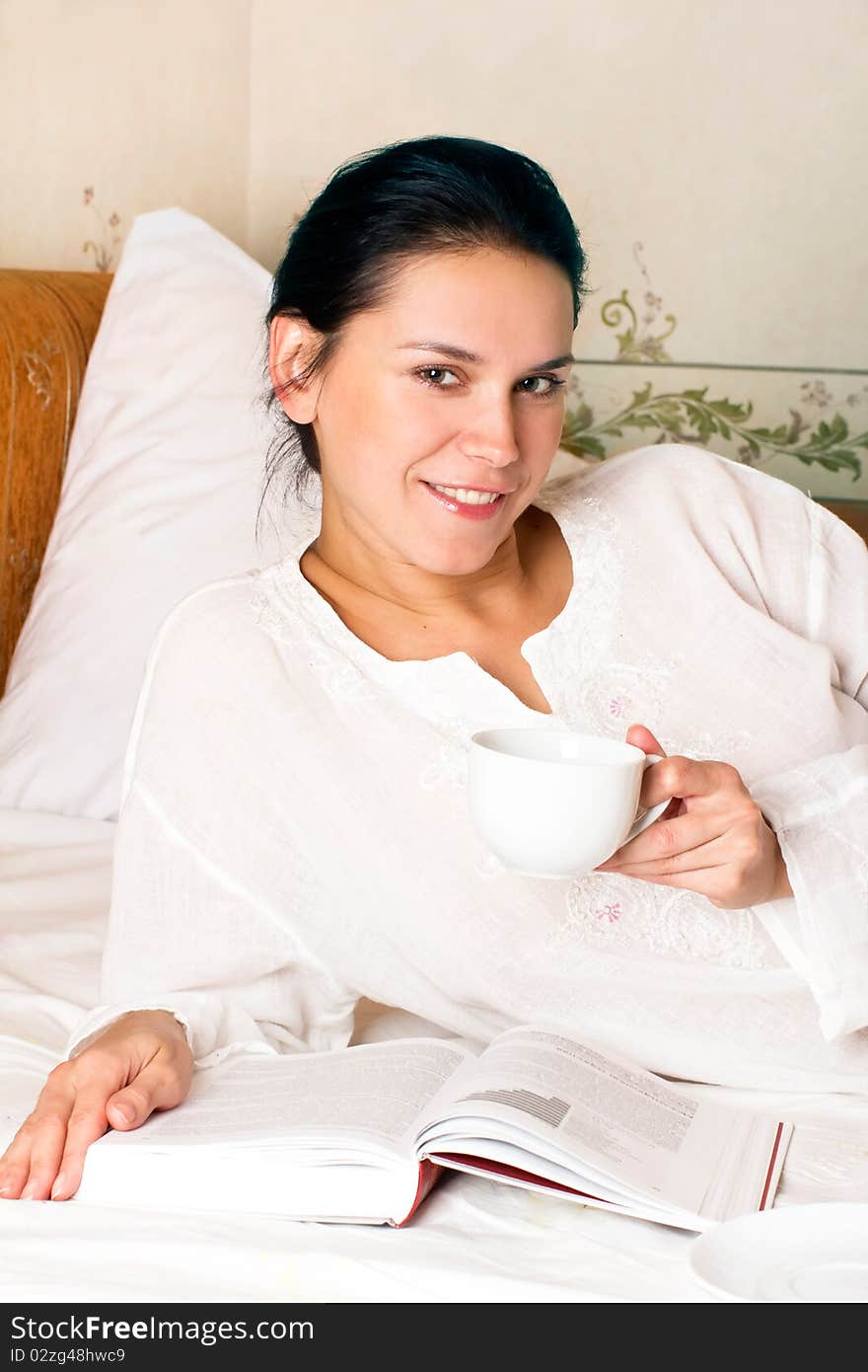 Portrait of young smiling woman with white cup and book on bed at bedroom