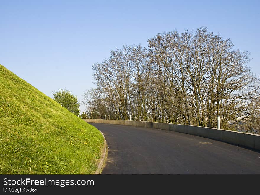 Road with a black coating in the park, the envelope of a hill with a growing spring grass on it. on the other side of the road there are trees and shrubs. Road with a black coating in the park, the envelope of a hill with a growing spring grass on it. on the other side of the road there are trees and shrubs