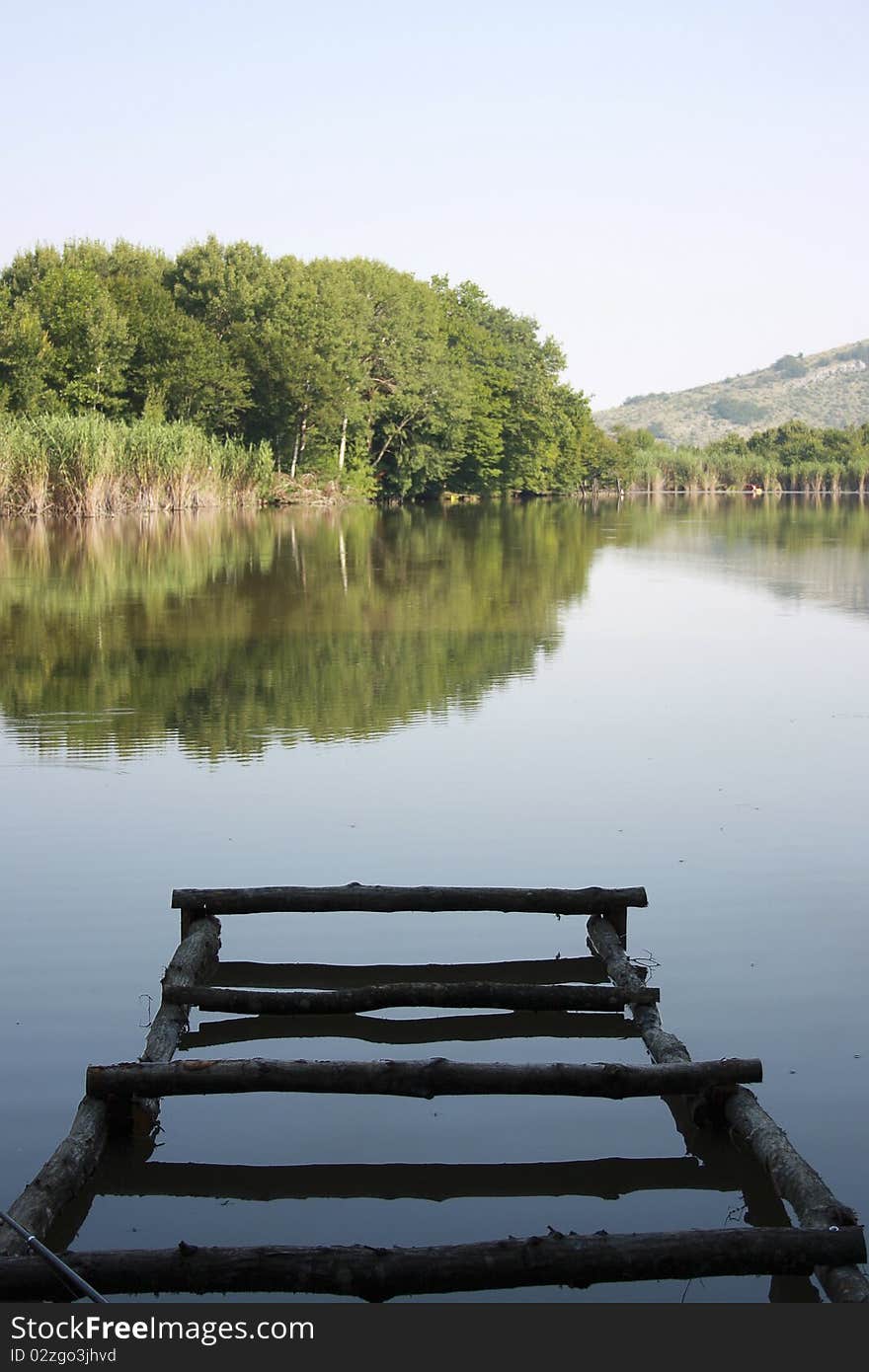 Smooth surface of lake in the morning, the shabby destroyed dock and the foreground, a cane and trees on lake coast on a background, the clear sky. Smooth surface of lake in the morning, the shabby destroyed dock and the foreground, a cane and trees on lake coast on a background, the clear sky