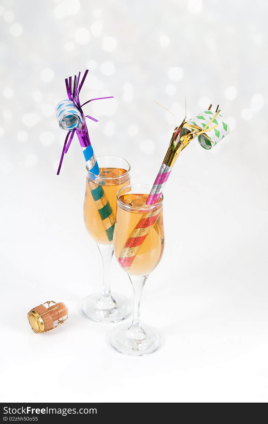 An angled studio shot of two New Year's Eve noisemakers in tow glasses of champagne with the cork on white with party lights in the background. An angled studio shot of two New Year's Eve noisemakers in tow glasses of champagne with the cork on white with party lights in the background.