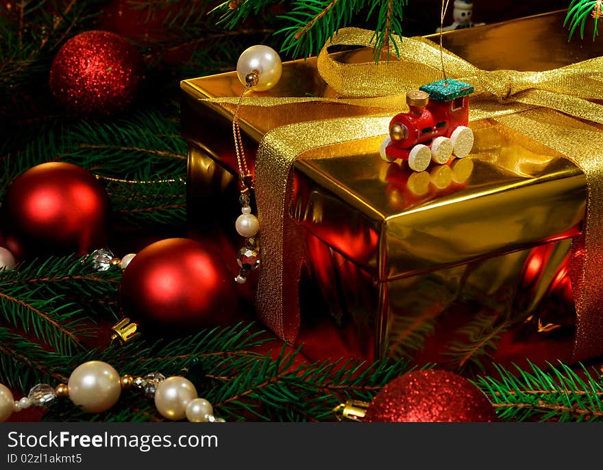 A gold box tied with gold ribbon surrounded by red christmas toys and christmas tree. A gold box tied with gold ribbon surrounded by red christmas toys and christmas tree