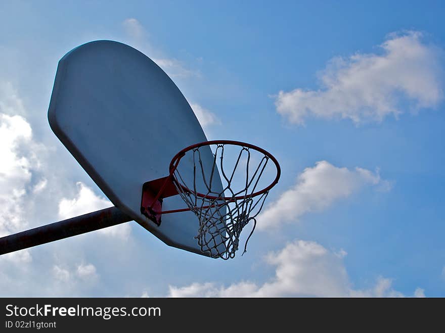 An empty basketball hoop, net swaying in the breeze, against a bright blue sky, with large clouds drifting from behind the backboard. An empty basketball hoop, net swaying in the breeze, against a bright blue sky, with large clouds drifting from behind the backboard.