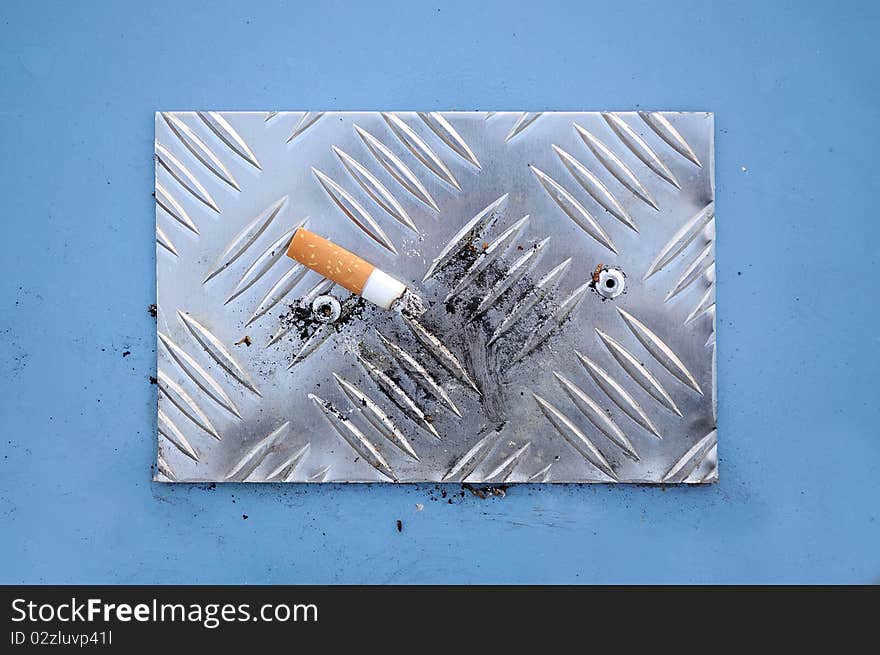 A cigarette end with ash on a metal cigarette stubbing plate on top of a litter bin. A cigarette end with ash on a metal cigarette stubbing plate on top of a litter bin