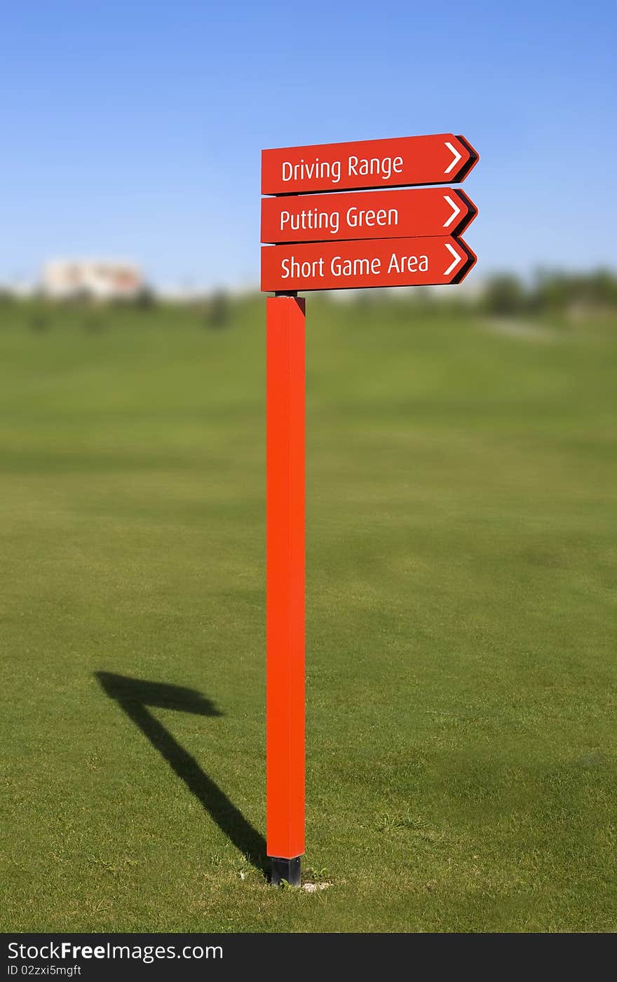 A red pole with sign arrows, pointing directions in a golf course: Driving Range, Putting Green, Short Game Area, over a clear blue sky with a blurred fairway on background. A red pole with sign arrows, pointing directions in a golf course: Driving Range, Putting Green, Short Game Area, over a clear blue sky with a blurred fairway on background