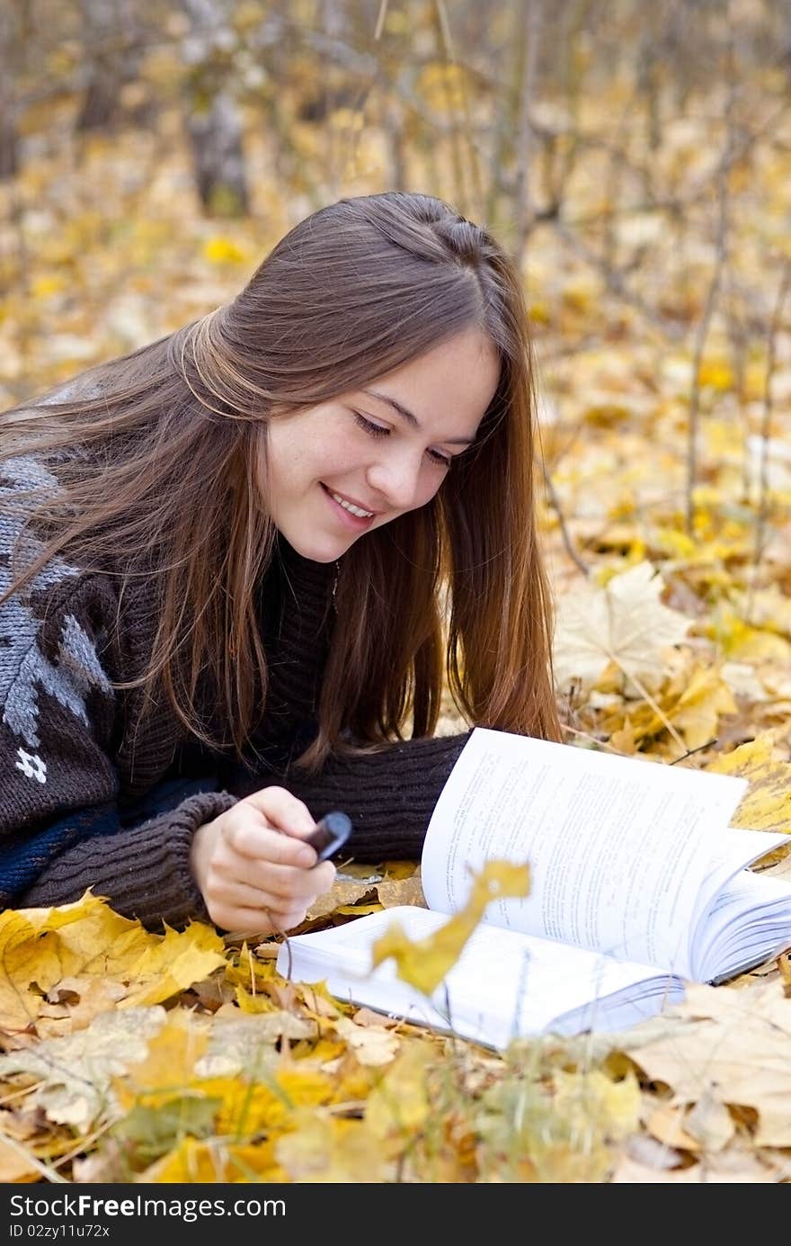 Portrait of smiling brown-haired girl in autumn park laying on leaves, reading a book. Portrait of smiling brown-haired girl in autumn park laying on leaves, reading a book