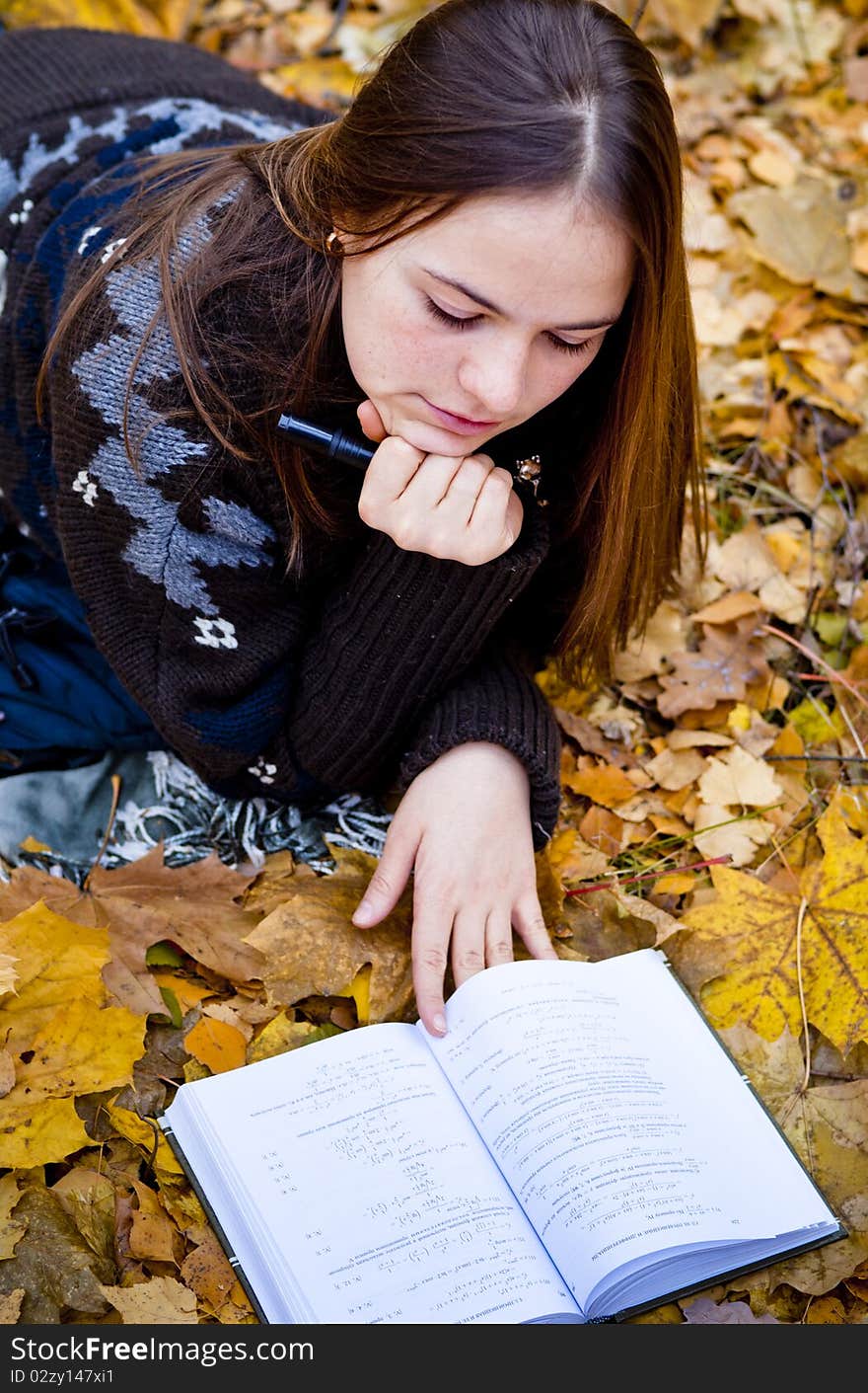 Portrait of thoughtful brown-haired girl in autumn park laying on leaves, reading a book. Portrait of thoughtful brown-haired girl in autumn park laying on leaves, reading a book