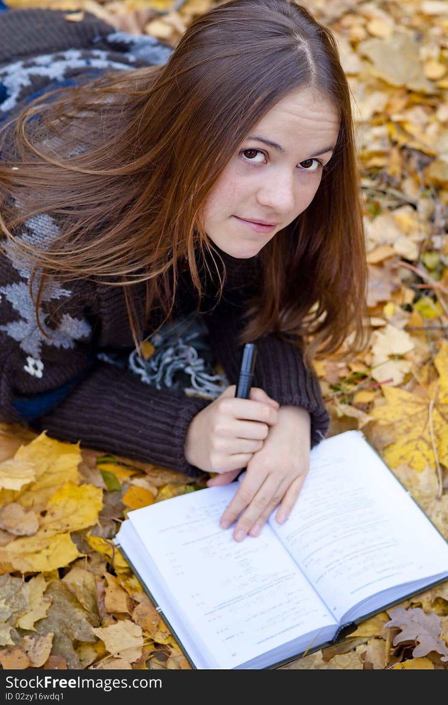 Portrait of brown-haired girl in autumn park laying on leaves, reading a book