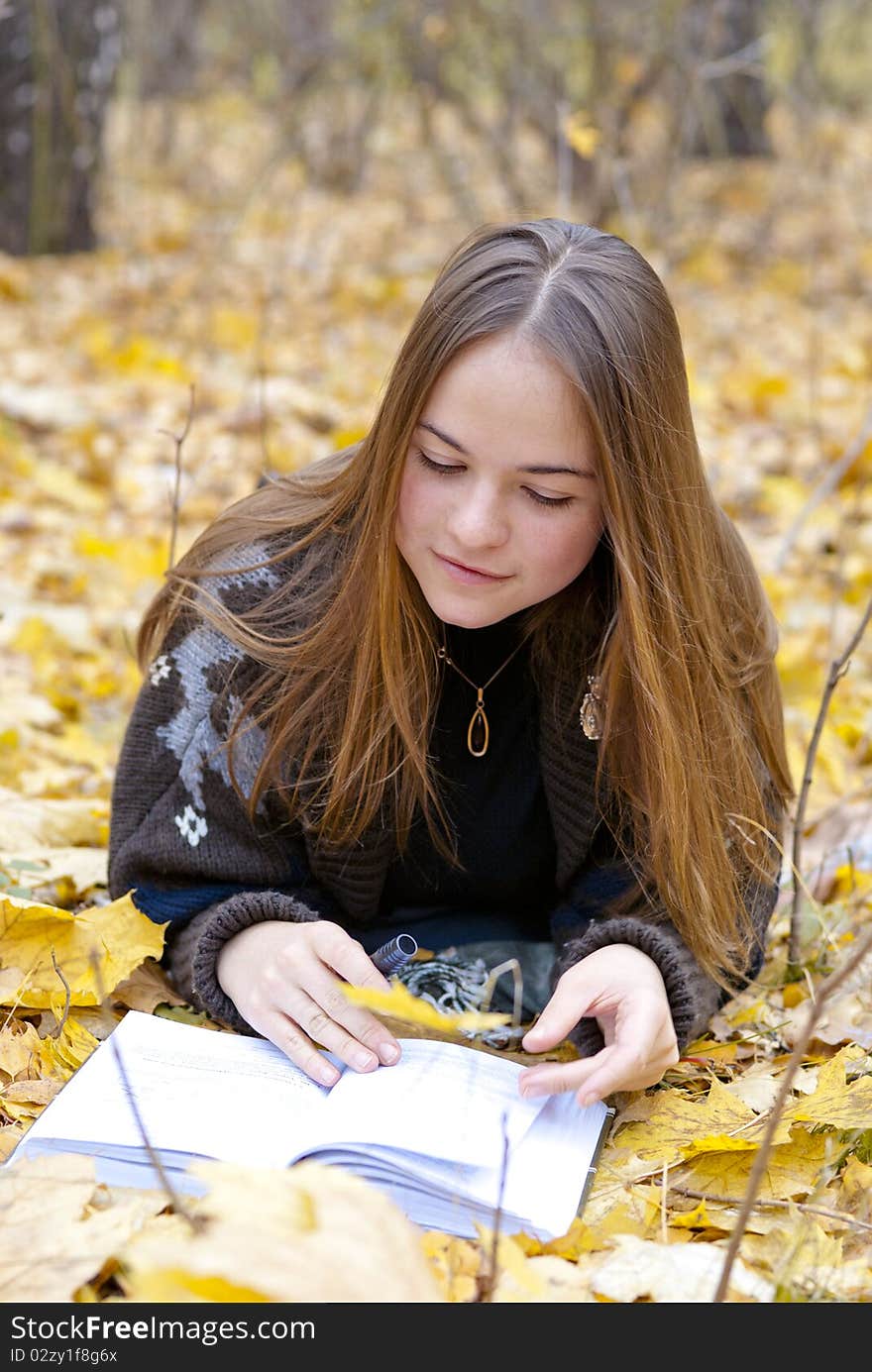 Portrait of brown-haired girl in autumn park laying on leaves, reading a book