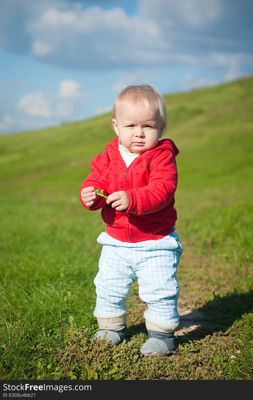 Adorable baby walking through green road within red jacket with hearts on mountain side. holding grass and looking forward. Adorable baby walking through green road within red jacket with hearts on mountain side. holding grass and looking forward