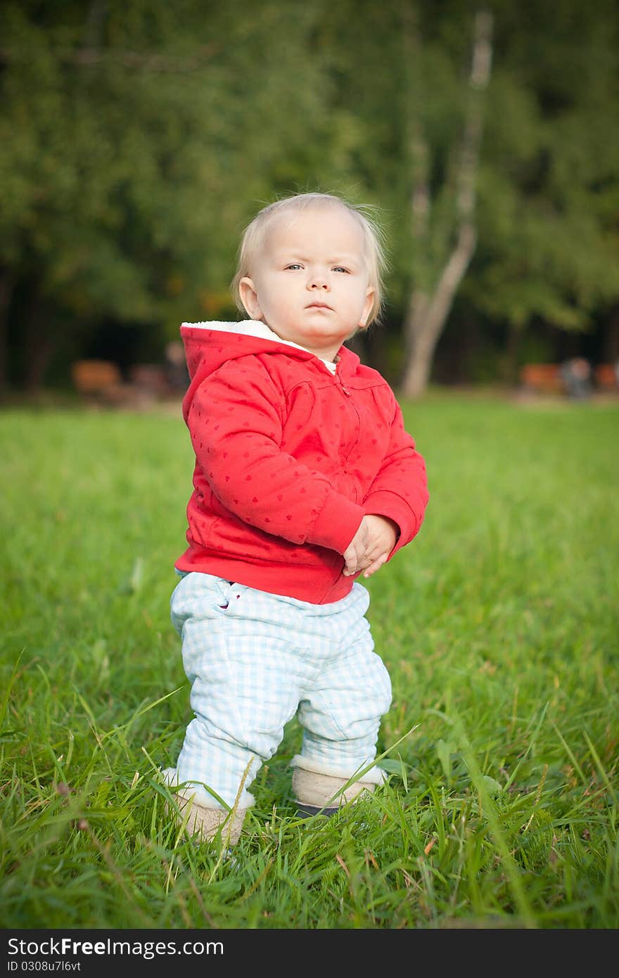 Adorable baby stay on green grass near forest within red jacket with hearts. looking forward