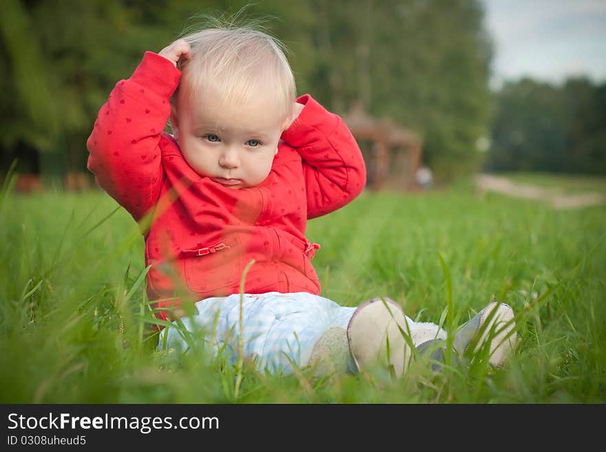 Adorable baby sit on green grass near forest within red jacket with hearts touching hair. looking forward. Adorable baby sit on green grass near forest within red jacket with hearts touching hair. looking forward