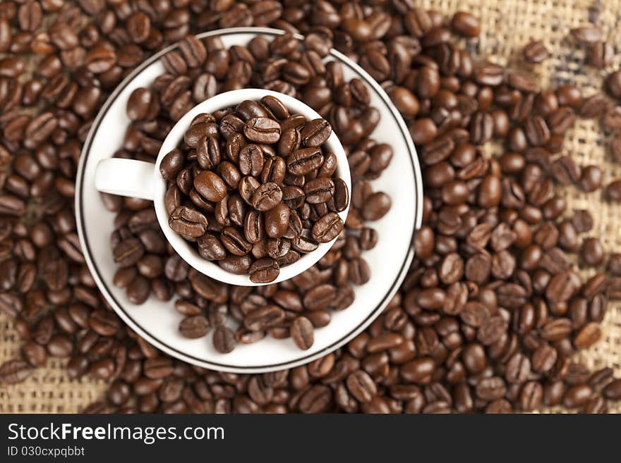 Espresso cup full with coffee beans. In background are more coffee beans visible.