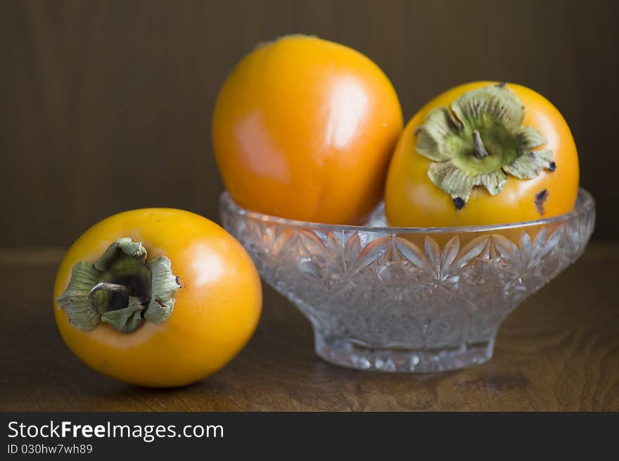 Still life of three Persimmons, two in a glass bowl. Everything decorated in front of dark brown background. Still life of three Persimmons, two in a glass bowl. Everything decorated in front of dark brown background.