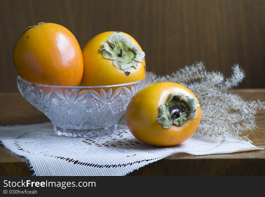 Three persimmons decorated in glass basin in front of dark brown wood background with a autumn grass. Everything is decorated on beige cloth. Three persimmons decorated in glass basin in front of dark brown wood background with a autumn grass. Everything is decorated on beige cloth.