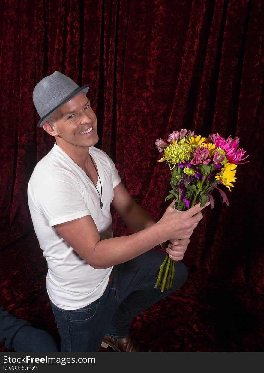 Smiling man wearing hat kneels with bouquet of flowers, photographed against red velvet background. Smiling man wearing hat kneels with bouquet of flowers, photographed against red velvet background