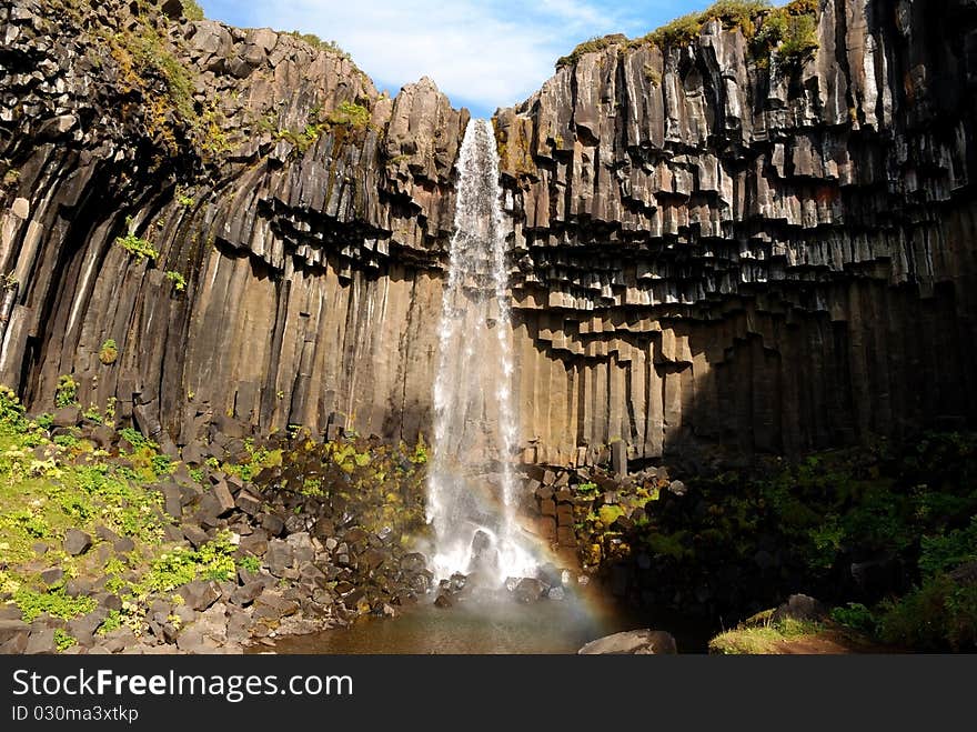 Narrow waterfall with basalt columns gallery forming small lake; little rainbow at the surface of the lake