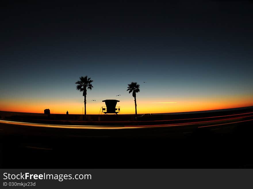 Silhouette of lifeguard tower at sunset, birds and airplane in the sky and car lights in motion. Silhouette of lifeguard tower at sunset, birds and airplane in the sky and car lights in motion.