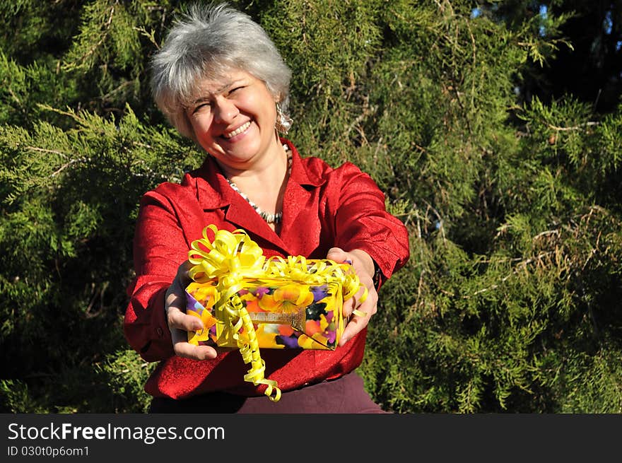 Lucky, an old woman holds out a gift in the yellow box, wrapped in gift paper and ribbons, against the backdrop of trees. Lucky, an old woman holds out a gift in the yellow box, wrapped in gift paper and ribbons, against the backdrop of trees