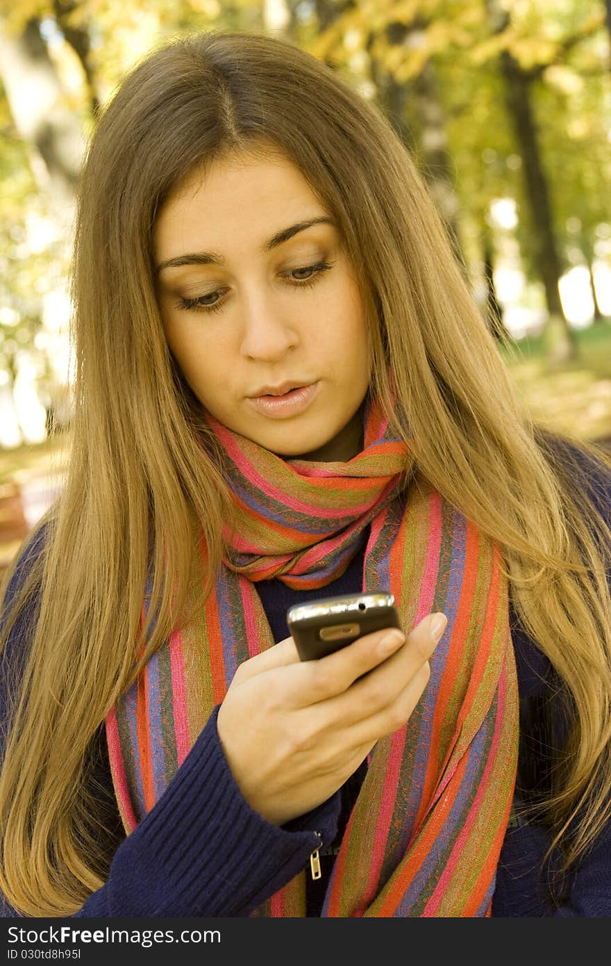 Young Caucasian woman calls on a mobile phone, sitting in a park on a wooden bench. Autumn around a lot of colorful foliage. Surprised. SMS. Young Caucasian woman calls on a mobile phone, sitting in a park on a wooden bench. Autumn around a lot of colorful foliage. Surprised. SMS