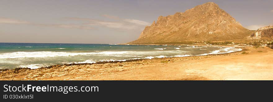 Long horizontal seaside panorama of a bay; rocky beach, sandy beach, resort village and hill in the distance Location: Cornino village, Gulf of Bonagia and view of Monte Cofano mountain in Sicily, Italy, Europe 3 horizontal images stitched together into a panoramic image