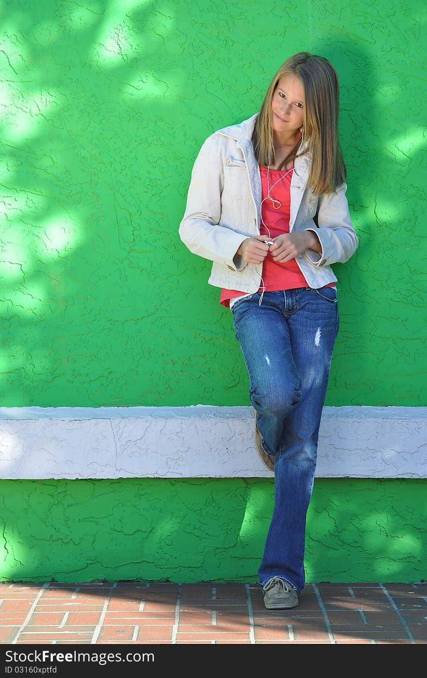 Pretty teen girl with long blonde hair wearing blue jeans leans against a green wall while she listens to tunes on her ear buds. Pretty teen girl with long blonde hair wearing blue jeans leans against a green wall while she listens to tunes on her ear buds.