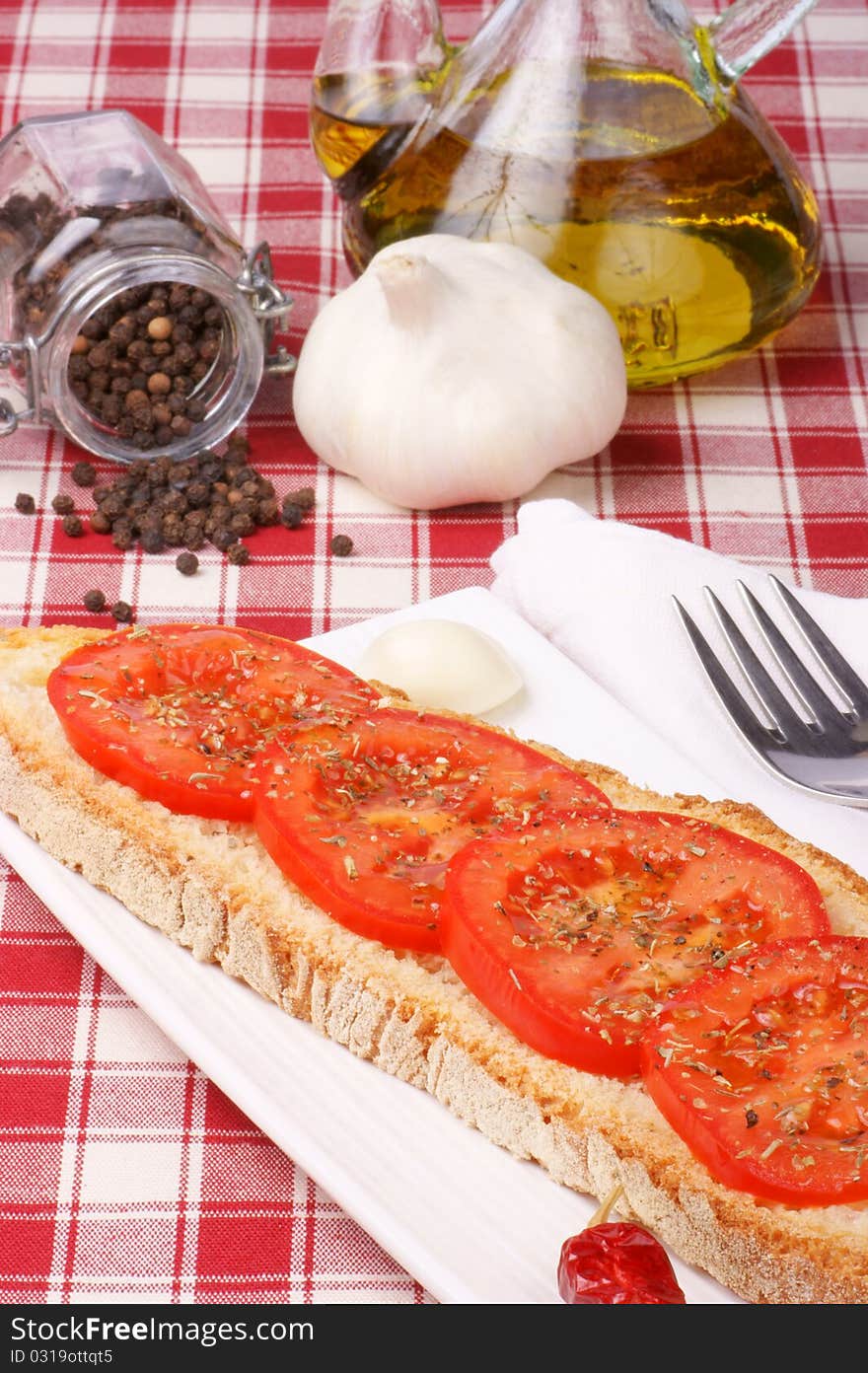 Toasted bread topped with tomato, olive oil and oregano served over a white plate with a garlic clove and a small red chilli pepper. Pepper, garlic and olive oil in the background. Toasted bread topped with tomato, olive oil and oregano served over a white plate with a garlic clove and a small red chilli pepper. Pepper, garlic and olive oil in the background.