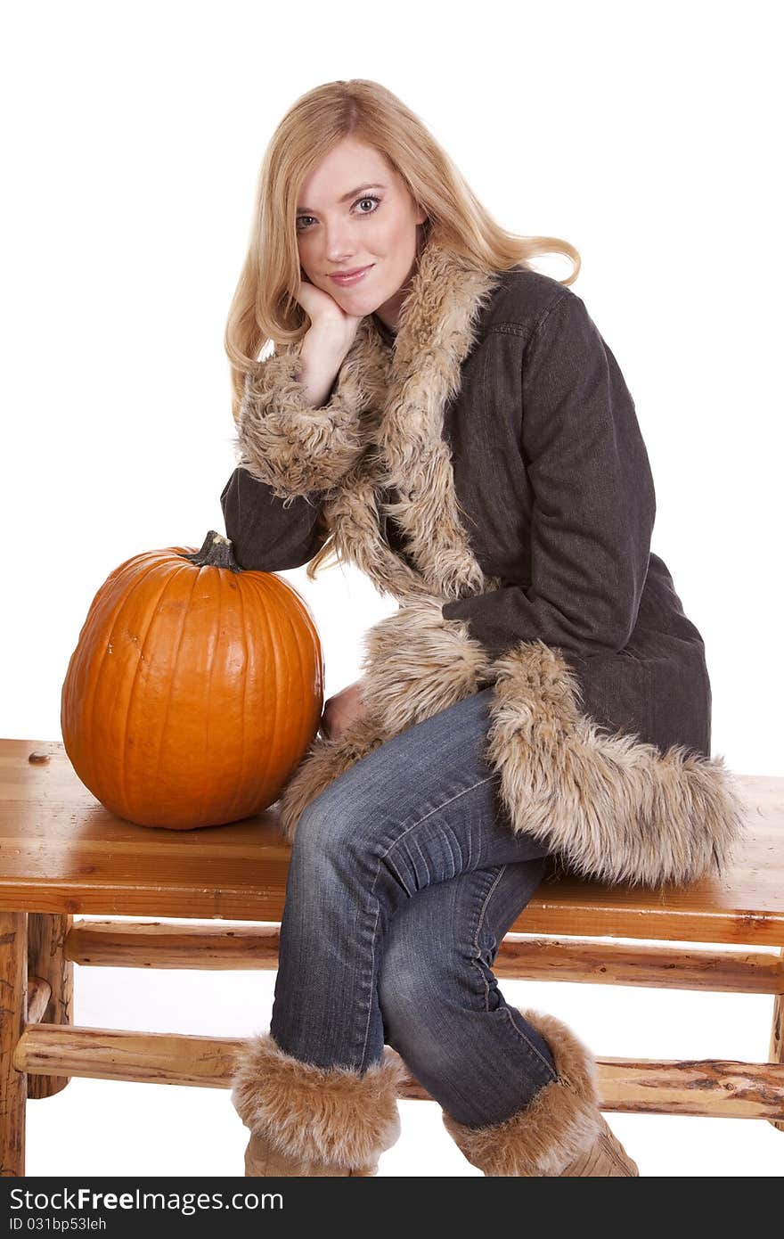 A woman sitting on a bench in her warm fuzzy coat while she leans on a pumpkin. A woman sitting on a bench in her warm fuzzy coat while she leans on a pumpkin.