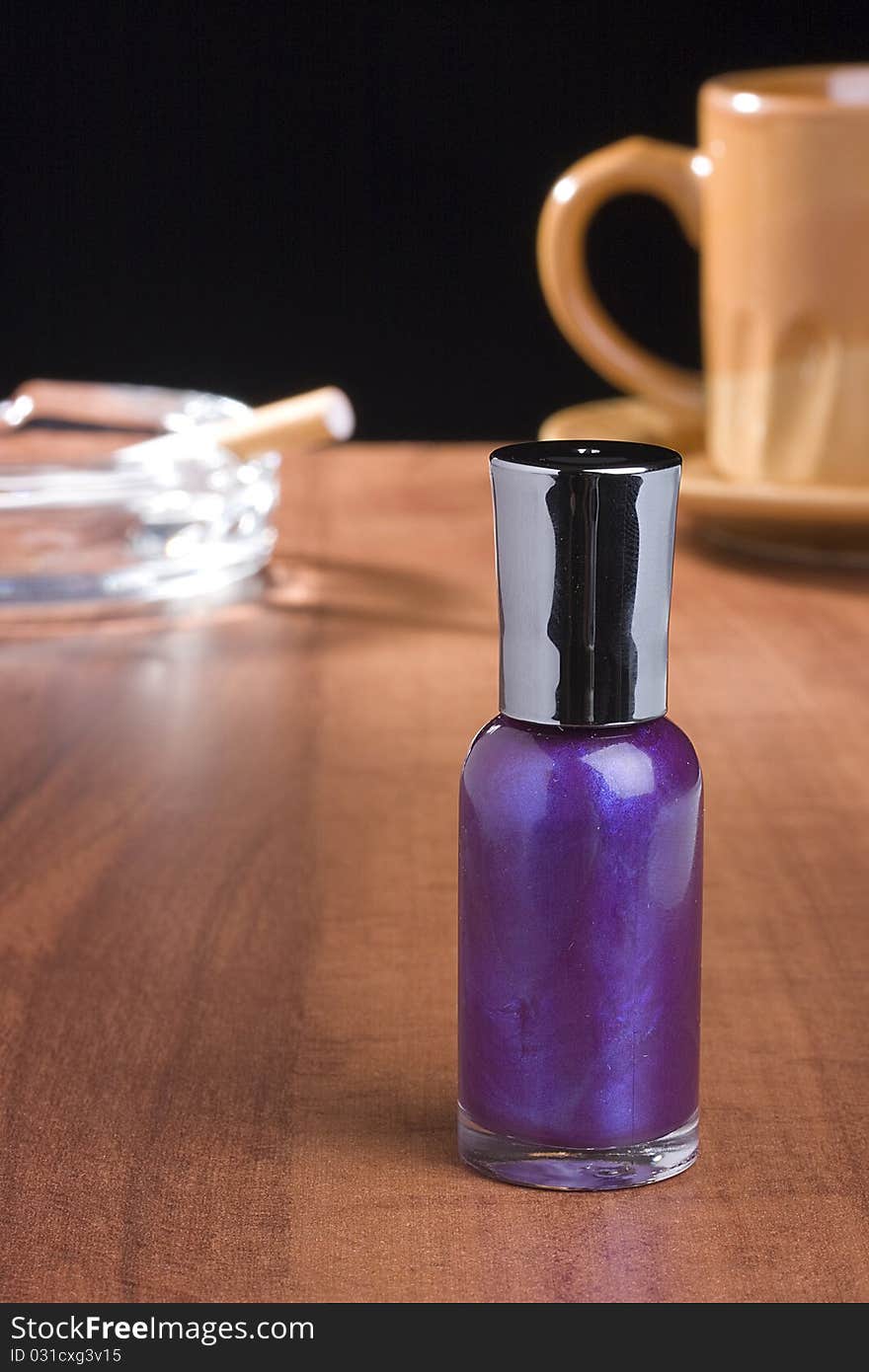 A bottle of purple nail polish standing on a coffee table in front of a cup of coffee and an ashtray.