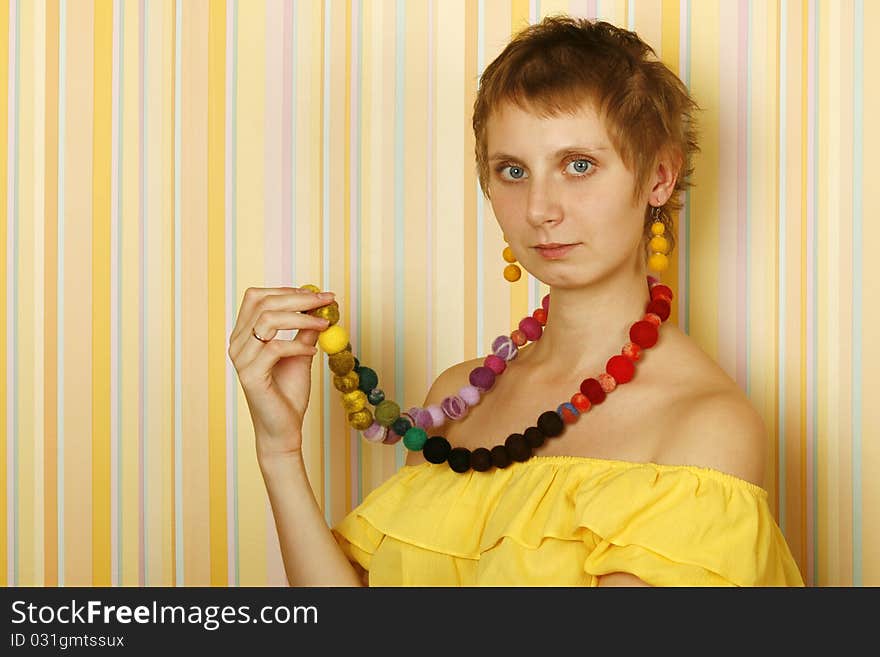 Young woman against yellow striped walls in a yellow blouse with bare shoulders in her wearing a necklace and earrings made of wool. Ethnos