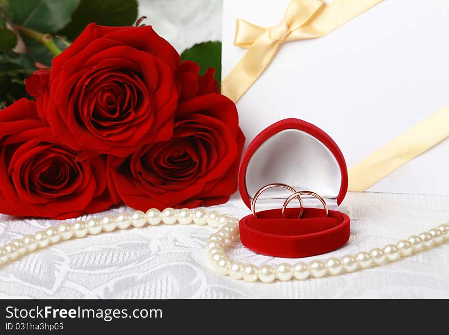 Wedding rings in box against bouquet of red roses and invitation card on lace background