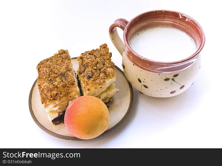 A cup of milk and a plate on which lay two pieces of cake and a ripe apricot, on a white background. A cup of milk and a plate on which lay two pieces of cake and a ripe apricot, on a white background